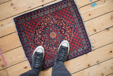 A person's perspective showing the scale of a vintage Kashan mat with a red and navy medallion.