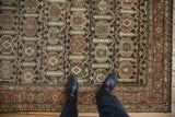 Distressed Hamadan rug's faded botanical pattern in charcoal, red, and beige, under black boots.
