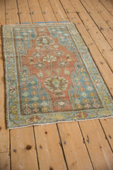 Angled view of a vintage Mazlaghan rug with a dusty red field and blue serrated corners.