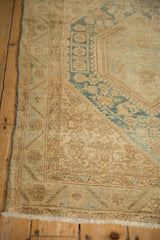 Distressed Sarouk rug corner showing worn beige pile, dusty blue floral patterns, and fringed edge.