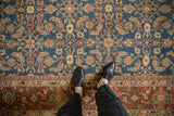 Feet on hand-knotted Afghani Mahal rug showcasing sapphire blue Herati field and rust Samovar border.