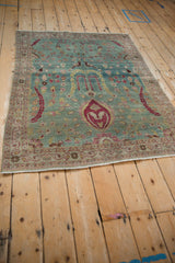 Angled view of a distressed Persian rug with birds and florals on a dusty blue-green ground.