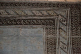 Corner of a distressed rug showing dark, multi-tiered borders against a worn, pale blue, hand-knotted field.