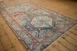Angled view of a distressed Malayer carpet with serrated medallions in dusty blue, cherry red, and sand.