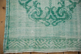 Distressed mint Oushak rug corner with emerald vinery, geometric border, and fringe.