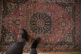 Persian Meshed rug's starburst medallion and floral vinery in cochineal red and navy blue, a person's feet.