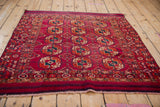 Angled perspective of a square Turkmen rug with a repeating geometric gul pattern in fuchsia and navy blue.