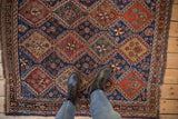 A person stands on a hand-knotted antique Qashqai rug, giving scale to its geometric blue and red panels.