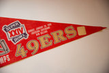 Angled close-up of red felt pennant tip, golden 49ERS lettering end, and tan patch.