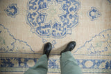 Distressed vintage Oushak carpet with beige field, pale blue arabesques, worn texture, and feet.