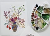 Floral watercolor print with blue vase and gold snake, next to a painter's palette.