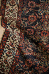 Rolled vintage Hamadan runner detail: worn dark navy Herati field, stacked henna border, tattered edge.