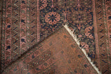 Folded Belouch rug corner reveals navy, dusty raspberry pile, woven foundation, and tattered fringe.