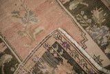 Distressed Oushak rug corner, dusty blush field, charcoal geometric border, and fringe.