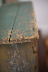 The weathered wooden corner of an antique stool with a heavily chipped turquoise paint patina.