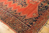 Corner angle view of antique tomato red Malayer rug with intricate dark blue border.
