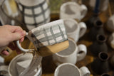 Hand holding speckled ceramic mug with hand painted black and white gingham and unglazed base.