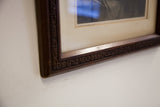 Corner detail of antique etching's hand-carved dark wooden frame with ornate scrolls.