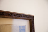 Close-up of antique etching's dark hand-carved wooden frame showing intricate scrollwork.