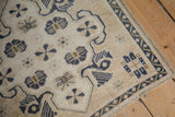 Corner detail of a distressed Oushak rug showing stylized floral motifs and a checkered border in navy and bronze.