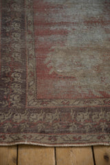 Distressed Oushak rug corner: worn dusty rose and grey pile revealing foundation, aubergine border.