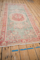 Distressed Oushak rug end, showing worn seafoam green pile, cherry red border, and fringe.