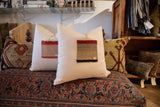 Cream pillow showcasing a textured Aguayo fragment with red, tan, black woven stripes and leather tag.
