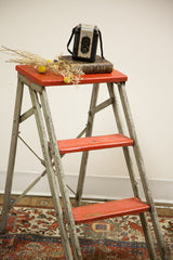 Close-up of industrial step stool's weathered silver legs and red ribbed steps over a patterned rug.