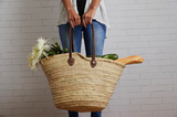 Woven Moroccan shopping basket with leather straps held by hands, filled with flowers and food.