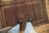 Belouch rug corner shows faded cochineal red, geometric lattice, exposed foundation, and tattered edge.