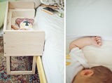 A modern white maple sidecar shown in use with a sleeping baby, placed next to a bed.