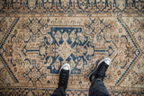 Distressed vintage Hamadan rug featuring a cobalt blue medallion and beige Herati pattern beneath sneakers.