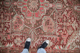 Feet in sneakers on a distressed vintage Heriz carpet, with geometric patterns, palmettes, and muted reds.