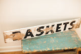 Chippy white paint, black 'ASKETS' letters, and exposed wood on the weathered sign.