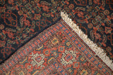 Folded rug corner reveals Herati pattern, navy marking on foundation, and cream fringe.
