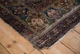 Antique Kermanshah rug corner with tattered edges, exposed foundation, and faded green gold arabesque border.