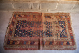 Heavily worn antique Kazak rug corner, faded ruby red, deep blue geometric designs, frayed fringe.
