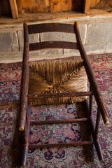 Aged dark wood and undone rush seat of an antique chair on a floral Kerman rug.