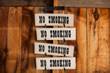 Four vintage No Smoking signs with black lettering on off-white plaques hanging vertically on a wood wall.