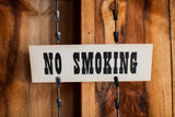 Close-up of vintage cream 'NO SMOKING' sign with bold black text and metal clips.