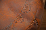 Antique leather pouf featuring dark orangey patina, embossed fan patterns, and thick texture.