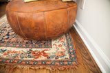 Patina of dark orangey leather pouf with embossed floral designs, on antique Bijar rug.