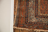 Antique Persian Serbend rug corner showing madder red and dark blue geometric borders with tattered fringe.