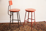 Adjustable metal legs of the industrial stool pair, one dark, one chippy red.