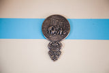 Ornate embossed metal art scene and scrolled handle on the back of the vintage handheld mirror.