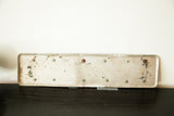 Back of vintage British license plate showing worn light grey metal with screws and rust.