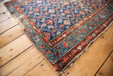Textured blue and red Hamadan rug corner, showcasing distressed Herati pattern and tattered fringe.