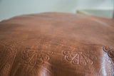 Natural brown leather pouf showing textured surface and embossed flower fan motifs.