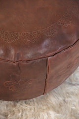 Distressed dark whiskey leather pouf with embossed fleur de lis and flower fan patterns.