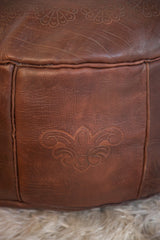 Distressed dark whiskey leather pouf close-up with fleur de lis and flower fan stenciling.